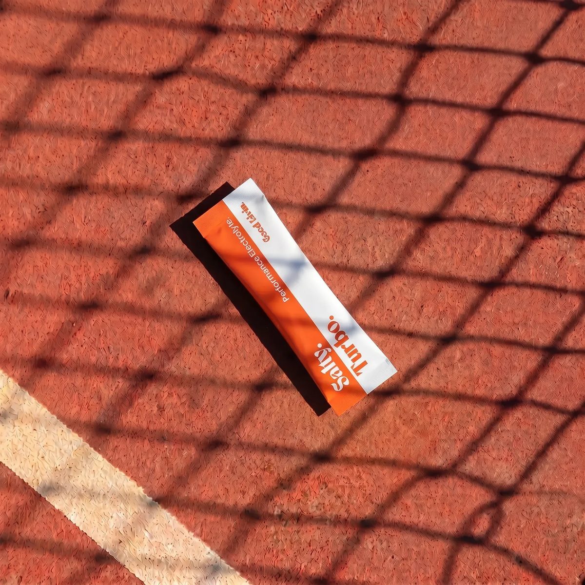 A single orange and white stick pack labeled 'Seltz' and 'Tart-Doo' lies on a red-orange textured surface with a black mesh net overlay casting shadows.